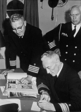 photograph of Captain O. C. Rohnke looking on as Admiral Richard E. Byrd writes a note to Karl E. Rohnke inviting him to join them on Operation Deepfreeze.  Other officer pictured is unidentified.  Circa 1955.